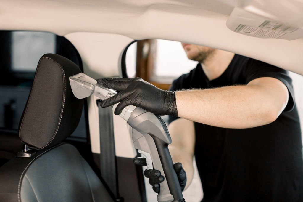 Young Caucasian man, car service worker, wearing black t-shirt, cleaning car front seat with wet vacuum cleaner. Car wet chemical vacuum cleaning, car detailing concept.