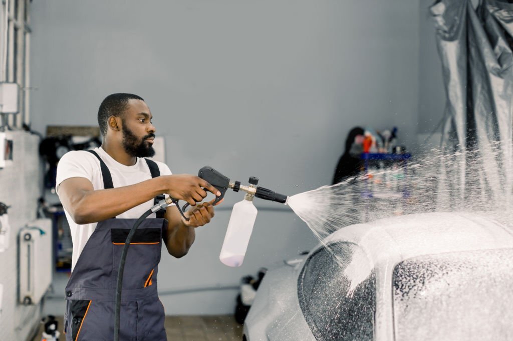 Close up portrait of young pleasant African car wash worker, wearing protective overalls, cleaning automobile with high pressure water jet at car wash, spraying the cleaning foam. Car wash detailing.
