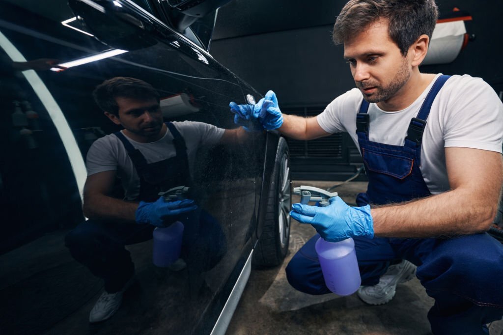 Automotive repairman in overalls using degreaser from spray bottle on car surface